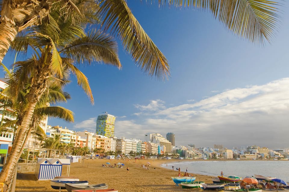 A beach with palm trees, boats, and buildings in the background.