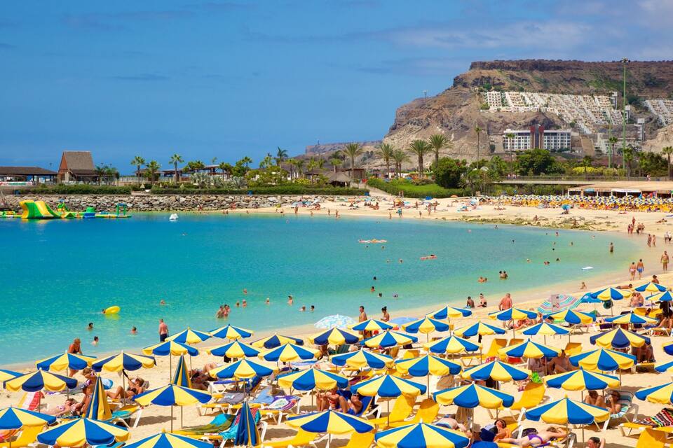 A crowded beach with blue and yellow umbrellas, clear blue water, and a rocky cliff in the background.