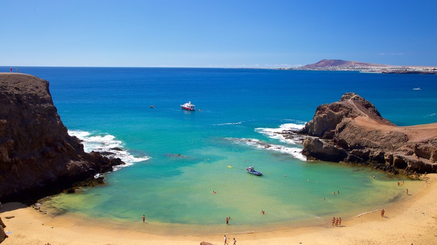 Una playa costera con aguas turquesas cristalinas, acantilados rocosos y una montaña distante.