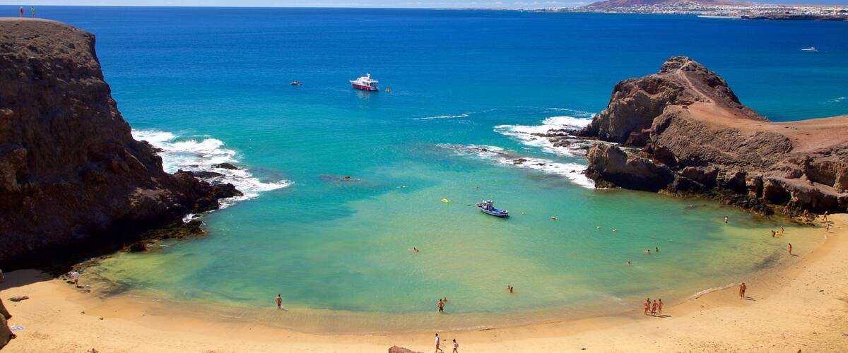 A coastal beach with clear turquoise waters, rocky cliffs, and a distant mountain.