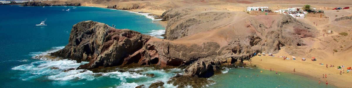 A coastal landscape with a rocky outcrop, clear turquoise waters, a sandy beach, and a mountain in the background.