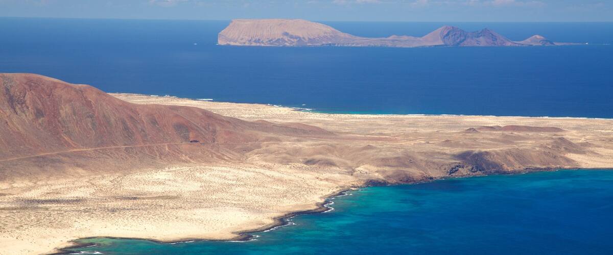A coastal landscape with a sandy beach, a rocky outcrop, and a distant mountain range.