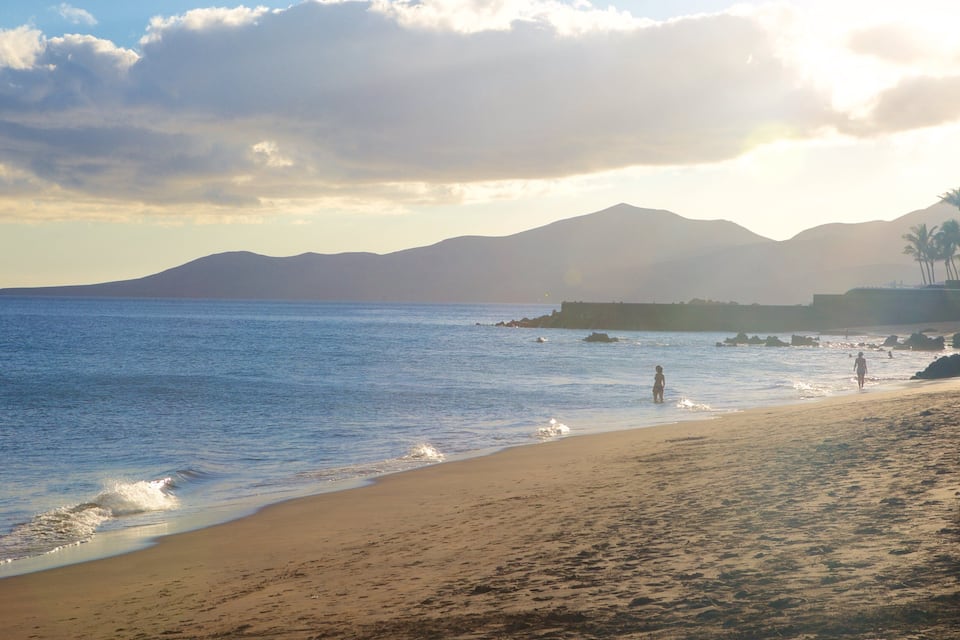 Una playa con aguas tranquilas, una orilla arenosa y montañas a lo lejos.