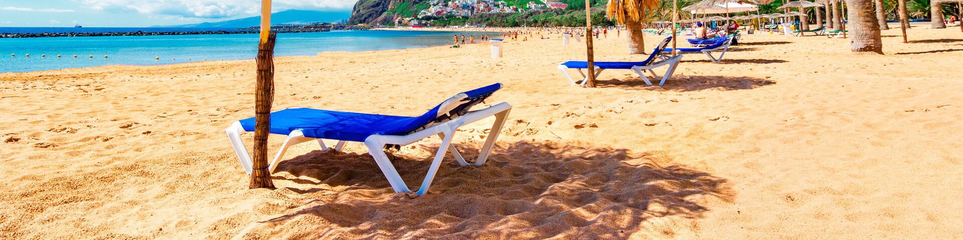 A beach with blue lounge chairs and straw umbrellas, a clear blue sky, and a mountain in the background.