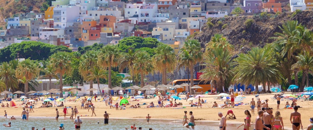 A crowded beach with people swimming and sunbathing, palm trees, and buildings in the background.