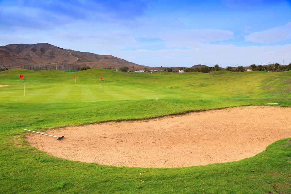 Un campo de golf con búnkeres de arena, banderas rojas y montañas al fondo.