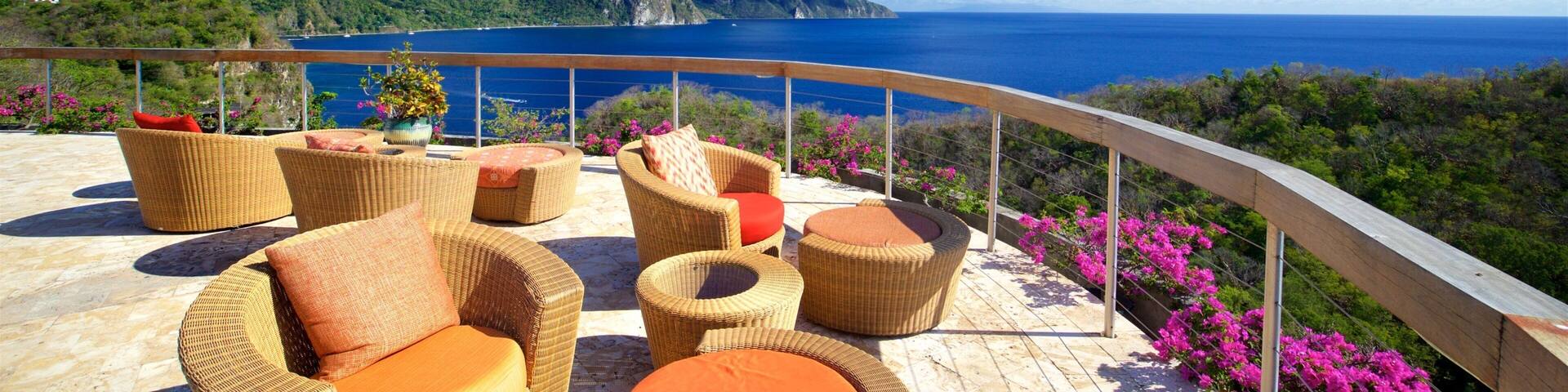 A terrace with wicker furniture overlooking a coastal landscape with mountains and the ocean.