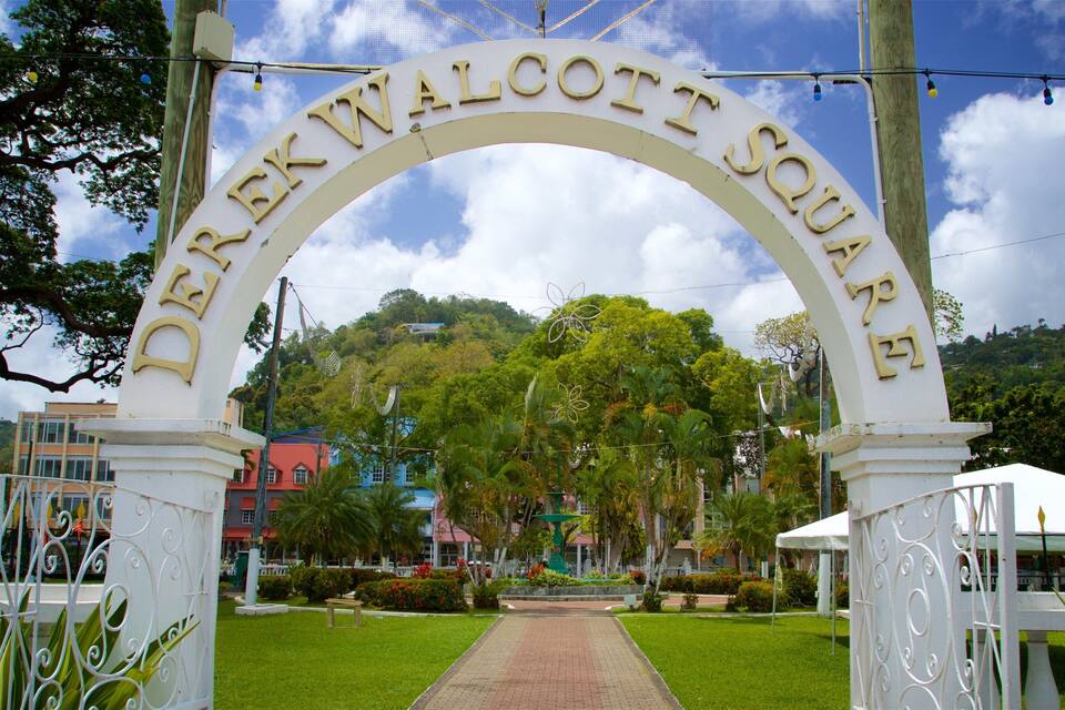 A white arched entrance with the text "DEREK WALCOTT SQUARE" against a backdrop of lush greenery and a clear sky.