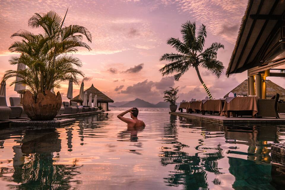 A man in a swimming pool at sunset with palm trees and thatched-roof huts.