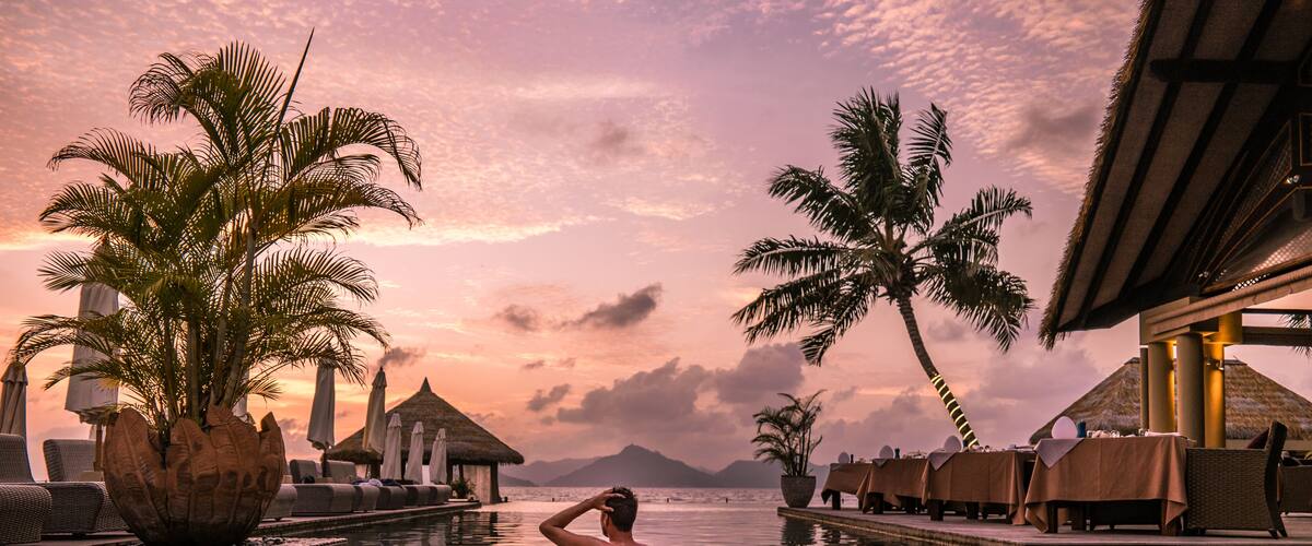 A man in a swimming pool at sunset with palm trees and thatched-roof huts.