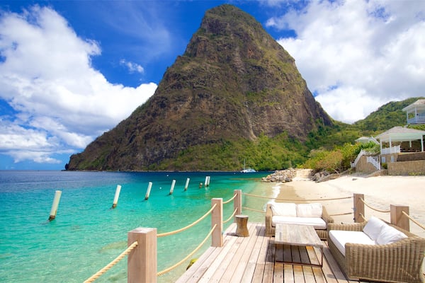 A wooden deck with wicker furniture overlooking turquoise waters, a prominent mountain in the background, and a white building with balconies.
