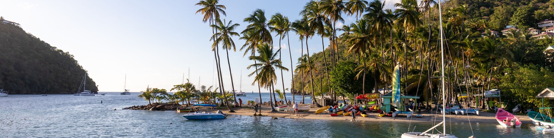 Cena costeira com céu claro, palmeiras, uma praia, barcos e uma paisagem montanhosa ao fundo.