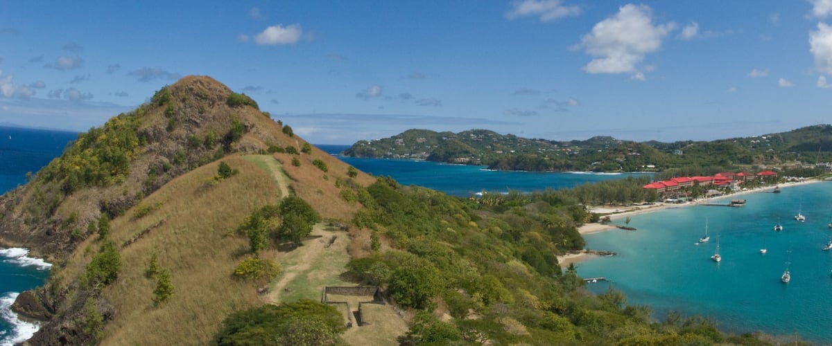 A coastal landscape with a prominent hill, clear blue sea, and a small settlement with red-roofed buildings.