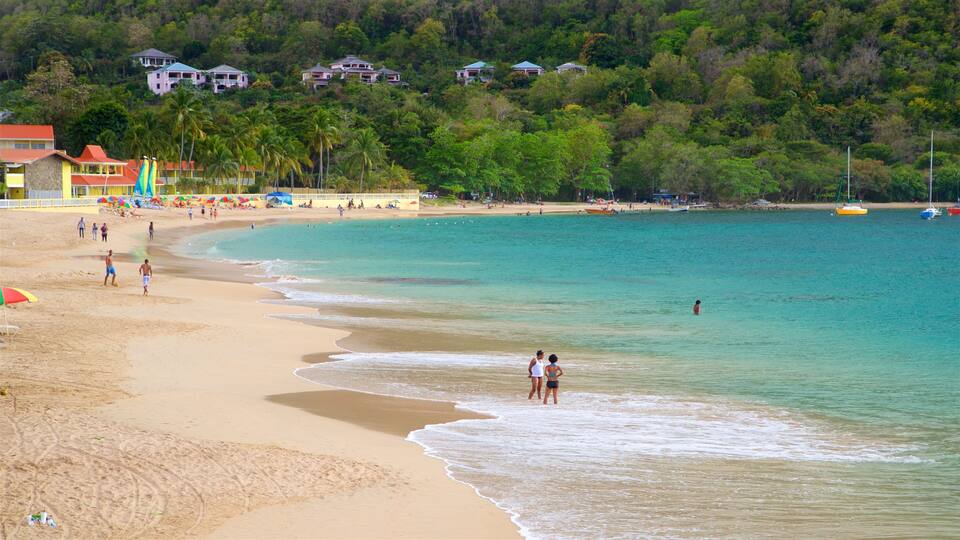 A beach with clear turquoise water, people walking along the shore, and houses on a hillside in the background.
