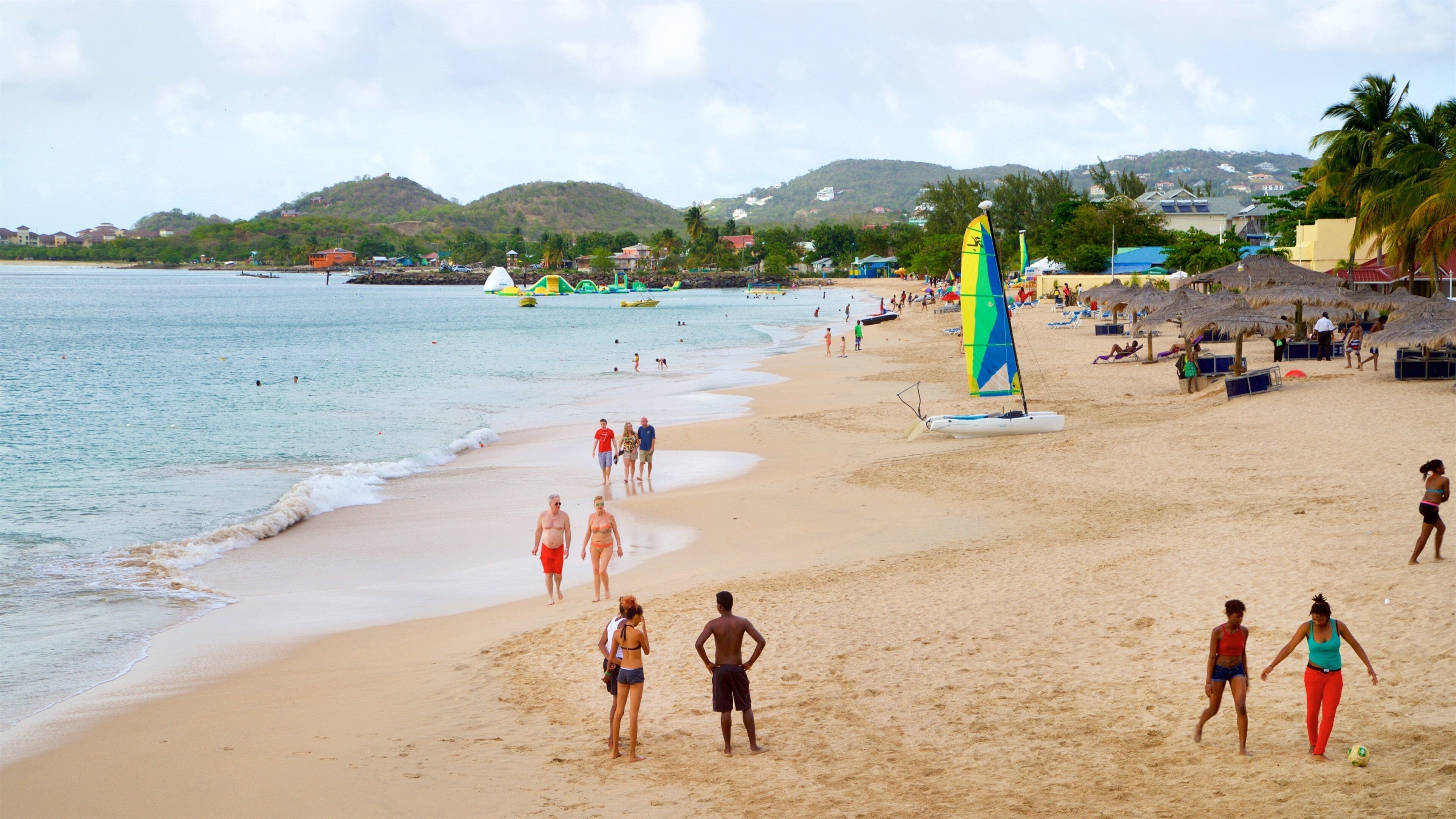 A beach with people walking, a sailboat, and a thatched-roof structure.
