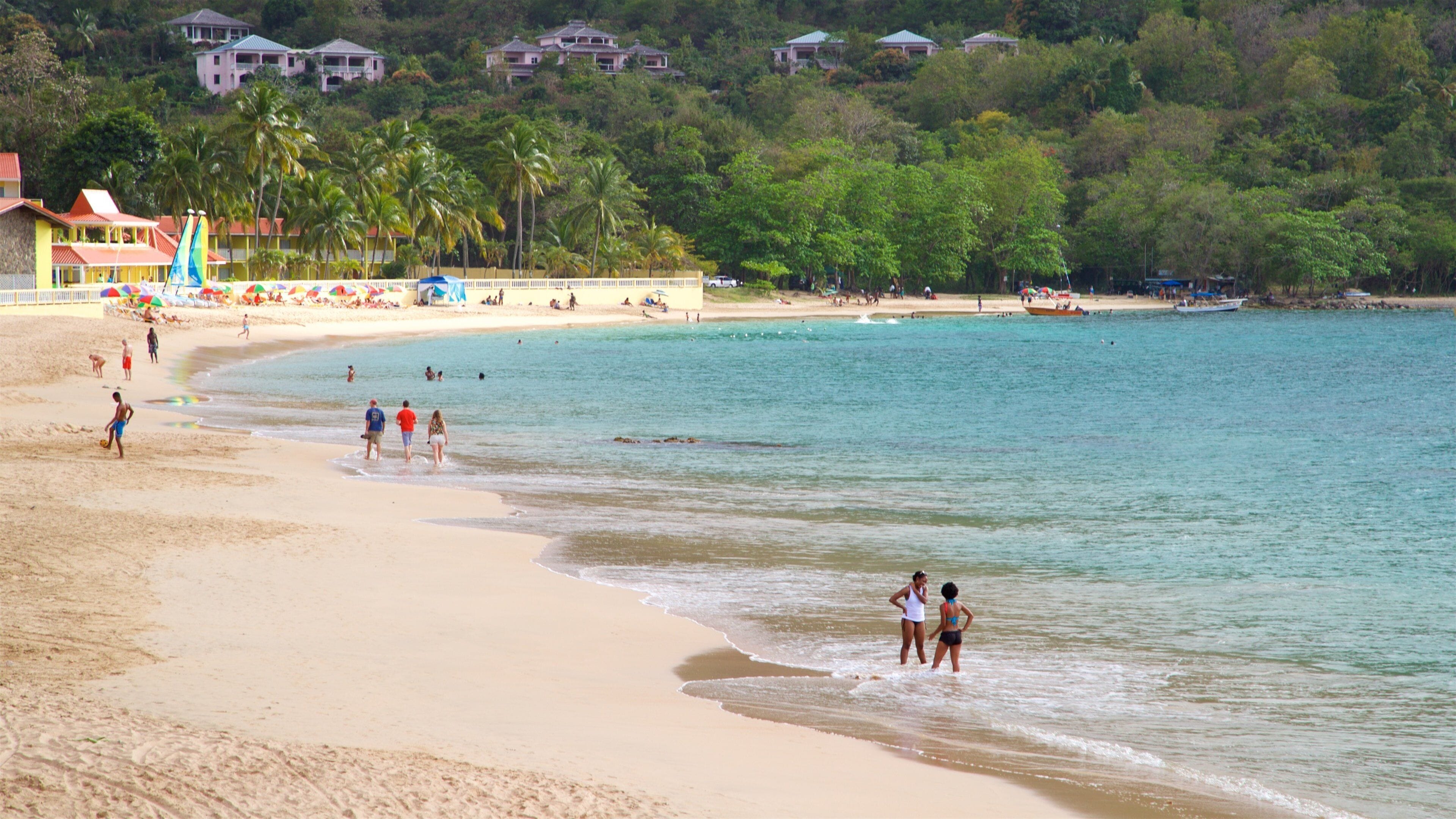 Una playa con gente caminando por la orilla, palmeras y casas al fondo.