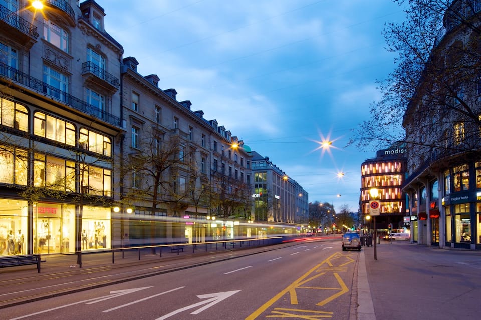 A city street at dusk with illuminated buildings, a tram, and a clear sky.