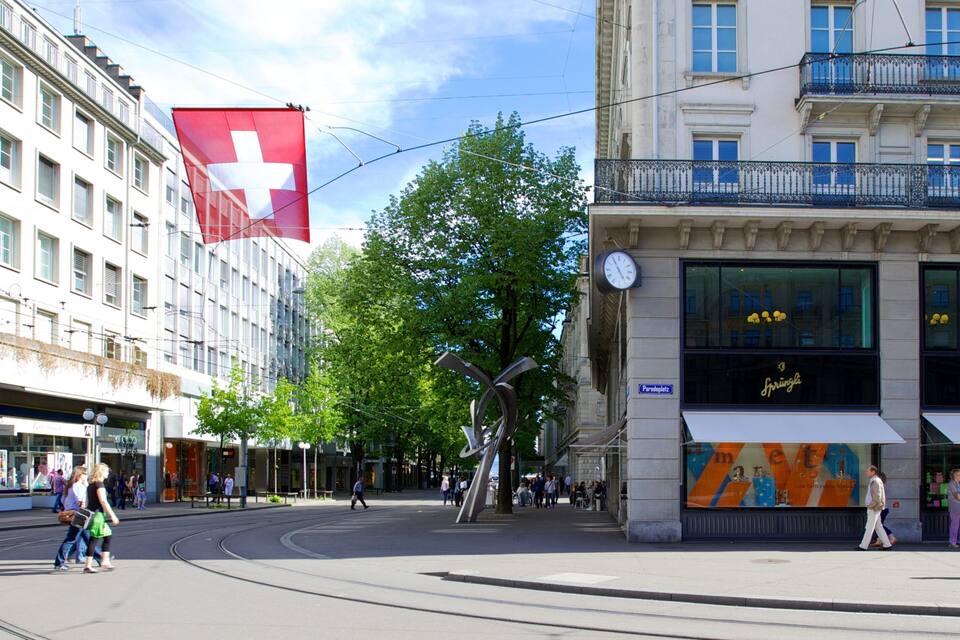 A city street with a large Swiss flag, a tram, and buildings with shops.
