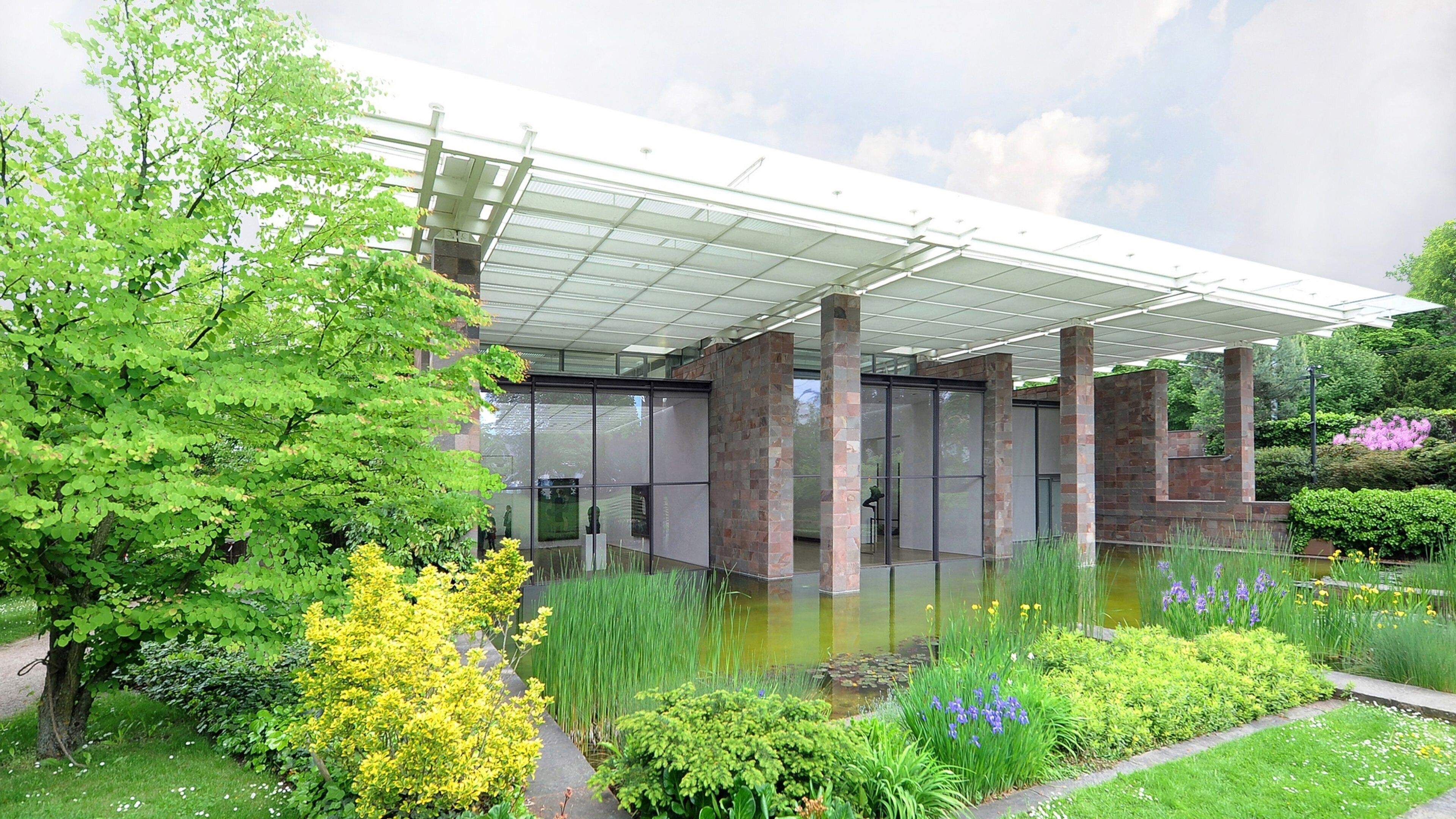 A modern building with a glass facade, surrounded by lush greenery and a small pond.