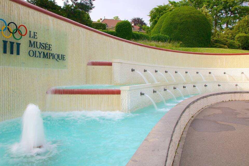A fountain with water jets, a sign for the Olympic Museum, and a landscaped area with trees and a hedge.