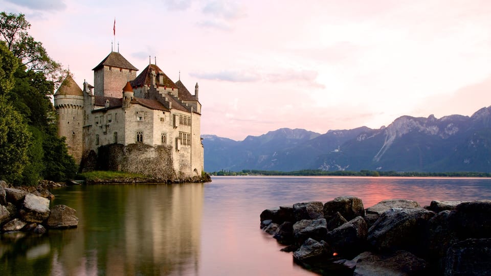 A castle by a calm lake with mountains in the background.