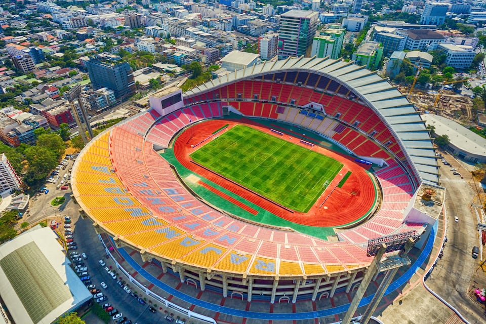A large, circular stadium with a green field and red seating areas.