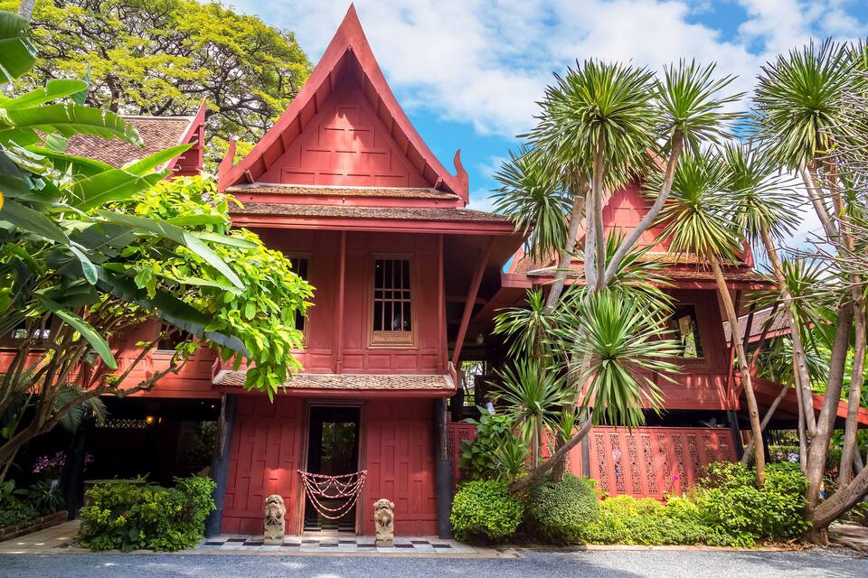 A traditional red-roofed building with a triangular roof and a small porch, surrounded by lush greenery and palm trees.
