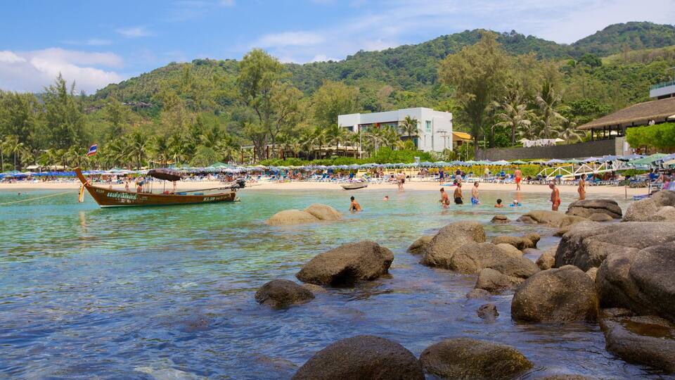 A beach with clear turquoise water, large rocks in the foreground, and a boat docked near the shore.