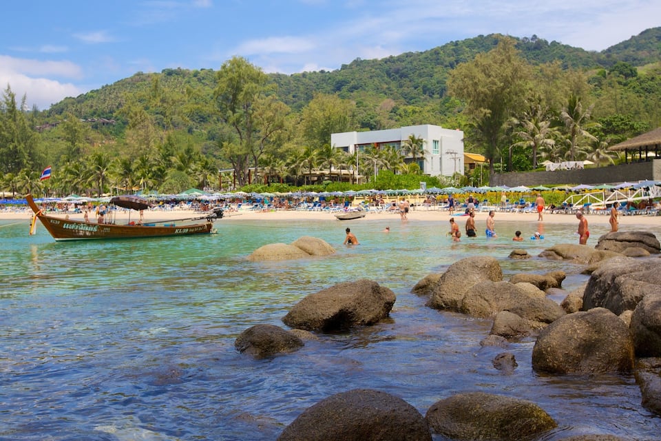 A beach with clear turquoise water, large rocks in the foreground, and a boat docked near the shore.