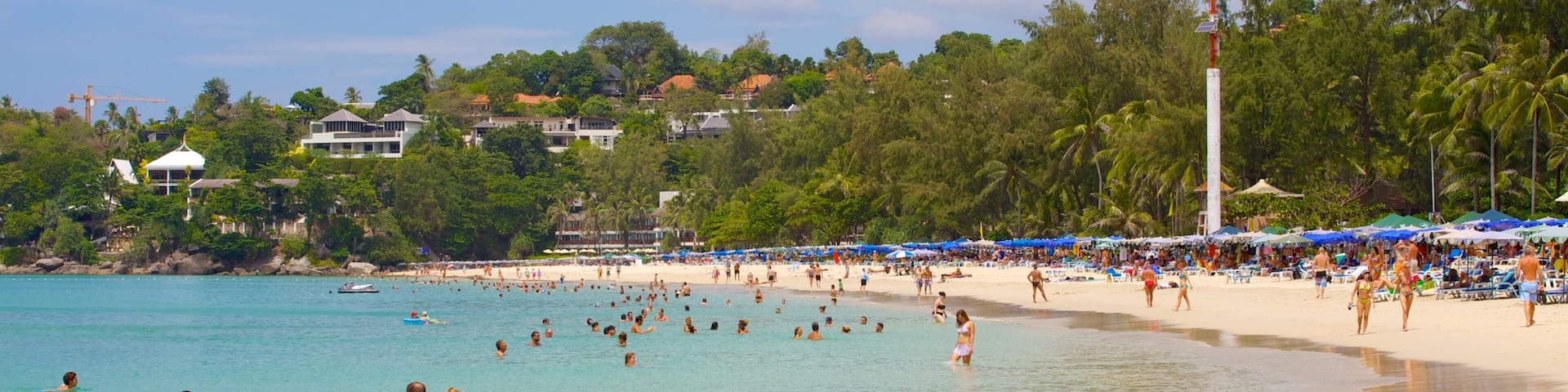 A crowded beach with clear turquoise water, palm trees, and beach umbrellas.