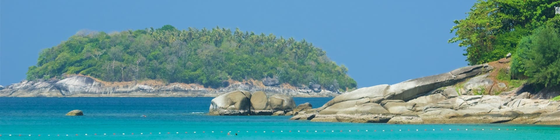 Mar turquesa y transparente con nadadores, una costa rocosa y una isla boscosa al fondo.