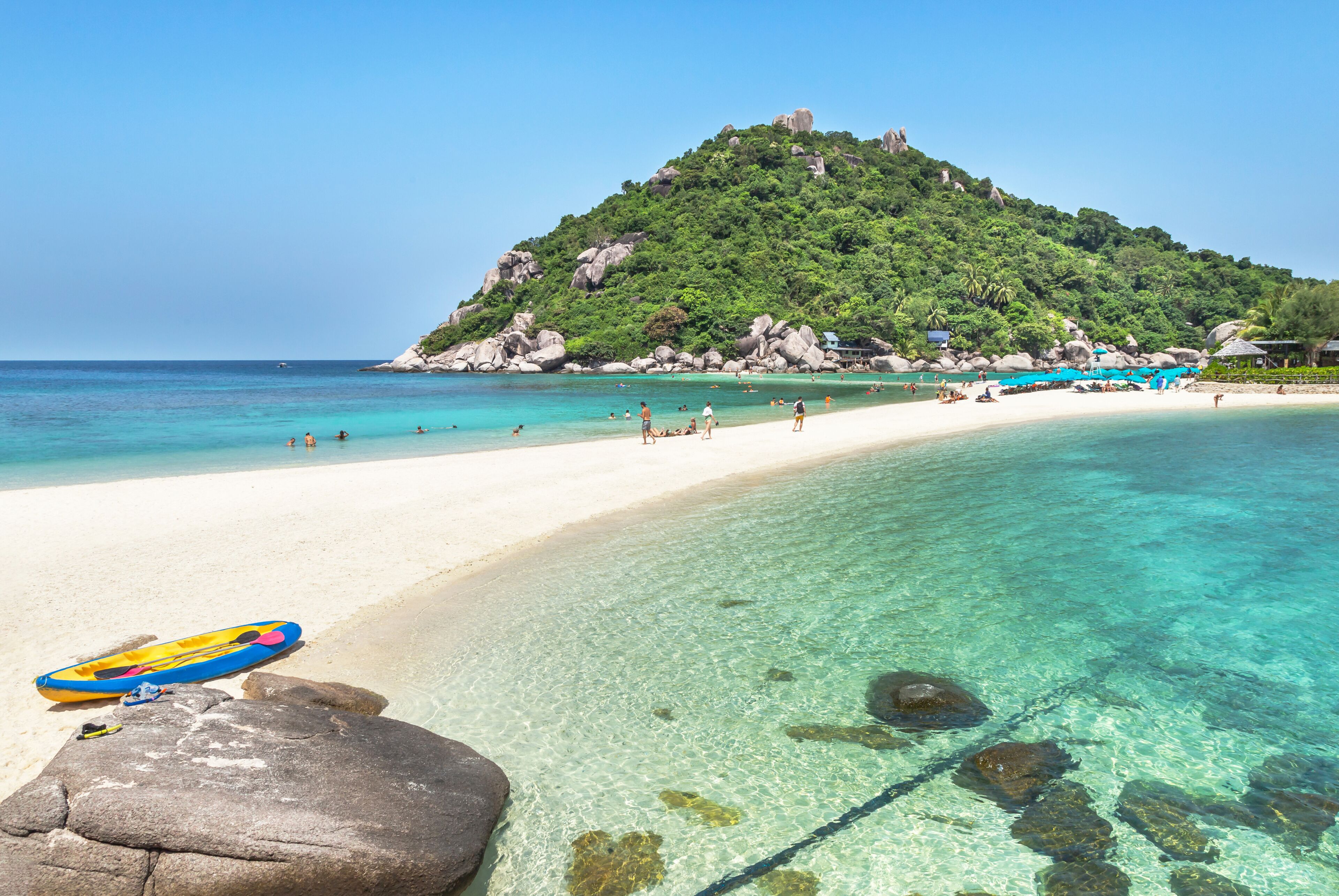 Uma praia com águas turquesa cristalinas, areia branca e uma colina verde com rochas.