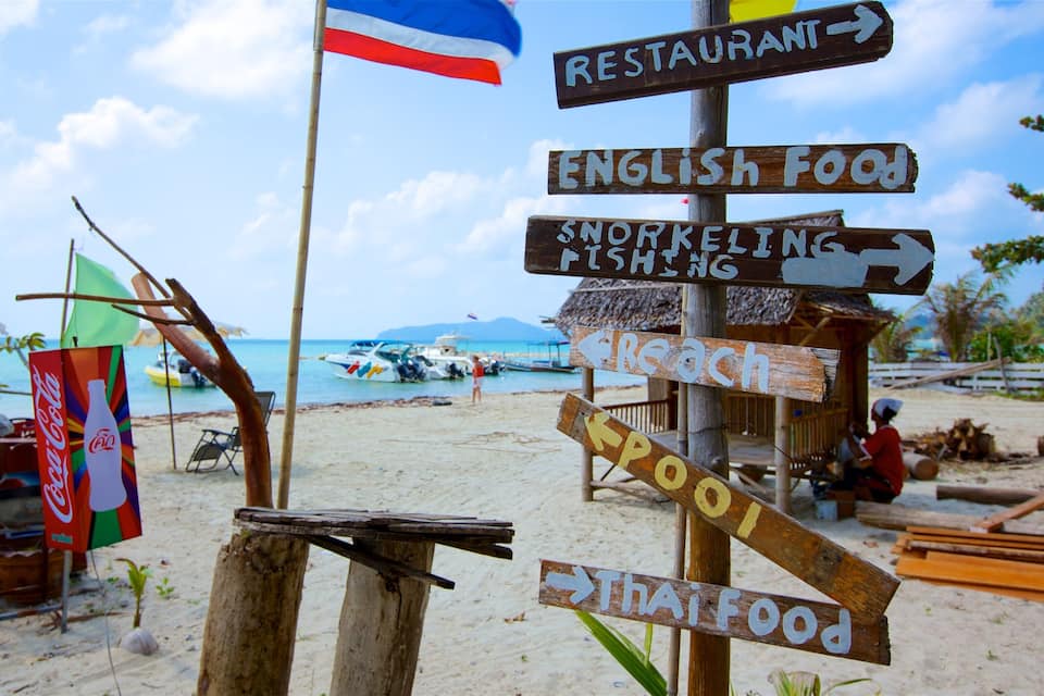 A beach with wooden signs pointing to various activities like 'Restaurant', 'English Food', 'Snorkeling', 'Fishing', 'Beach', 'Pool', and 'Thai Food'.