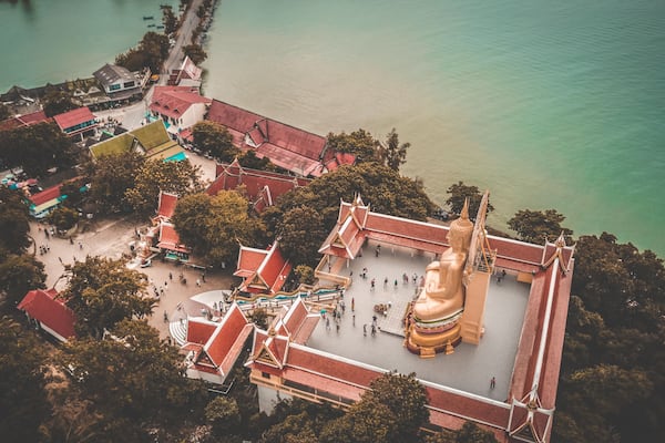 An aerial view of a temple complex with a large golden statue, surrounded by traditional architecture and a calm body of water.