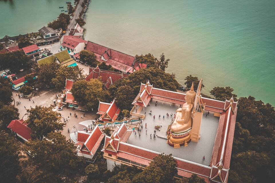 An aerial view of a temple complex with a large golden statue, surrounded by traditional architecture and a calm body of water.