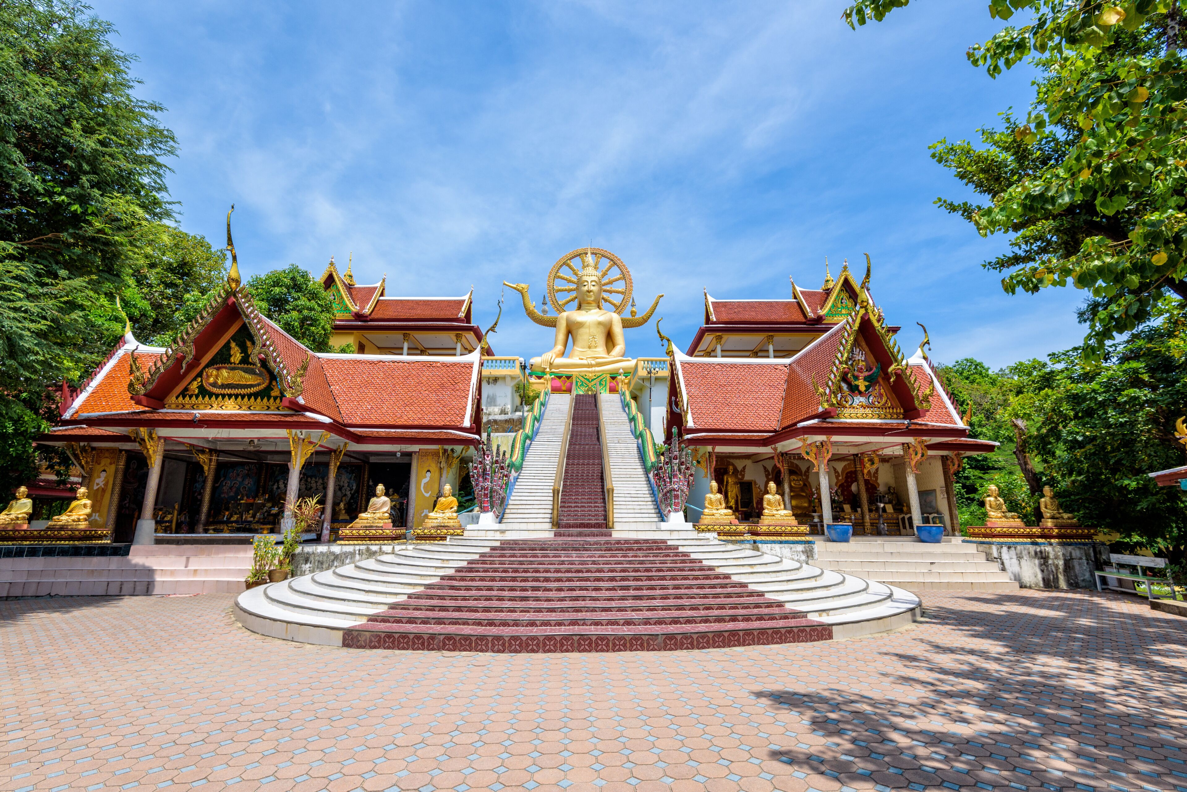 A temple with a large golden statue, red-roofed buildings, and a staircase leading to the statue.