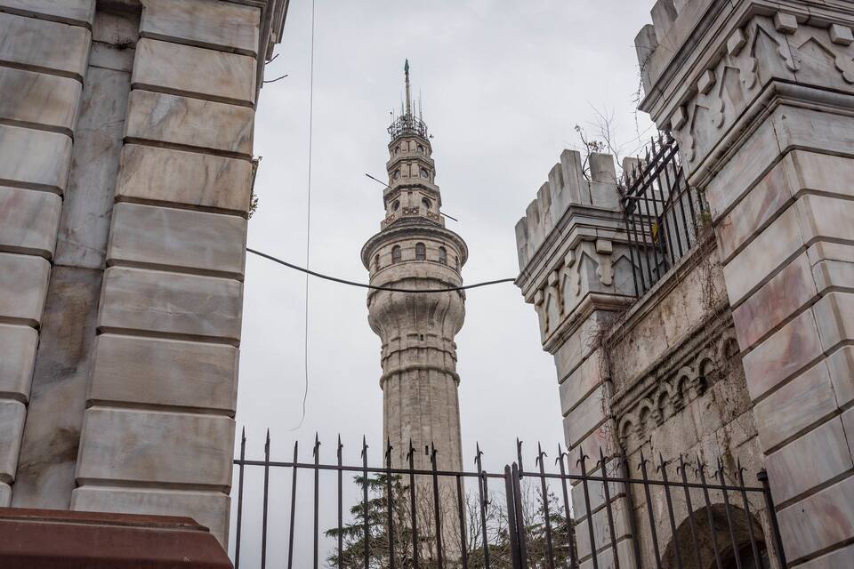 Uma torre histórica com trabalhos em pedra detalhados e uma cerca de metal ao primeiro plano.