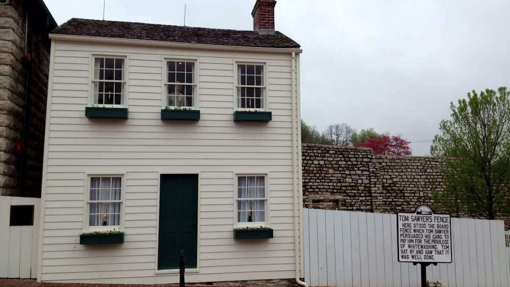 Un bâtiment blanc avec des encadrements de fenêtres verts et une porte verte, un mur de pierre et une pancarte concernant la clôture de Tom Sawyer.