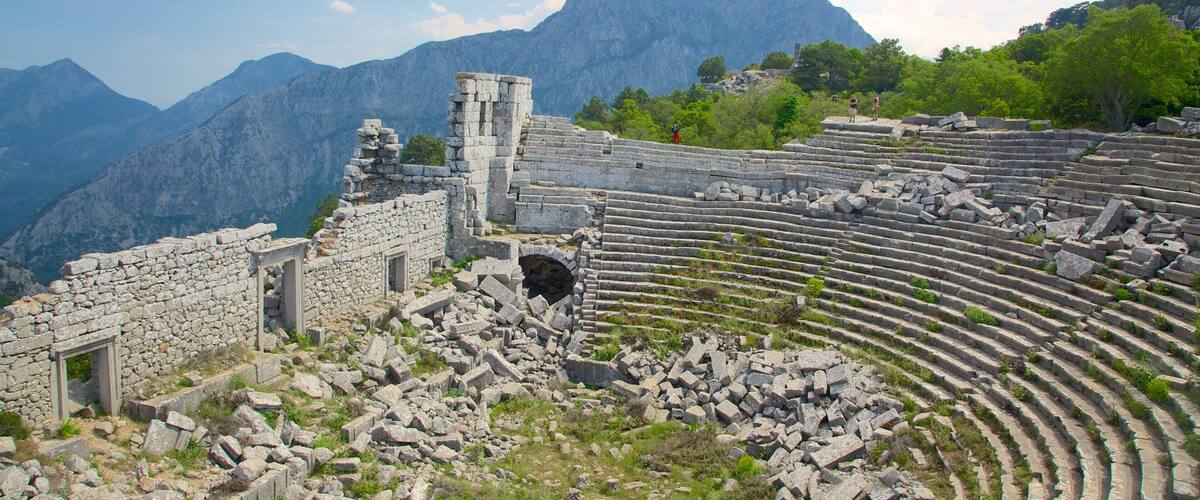 Ancient ruins with stone steps and a partially intact structure, set against a backdrop of mountains and lush greenery.