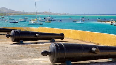 Une scène côtière avec plusieurs bateaux sur une eau turquoise, un mur jaune et deux canons noirs.