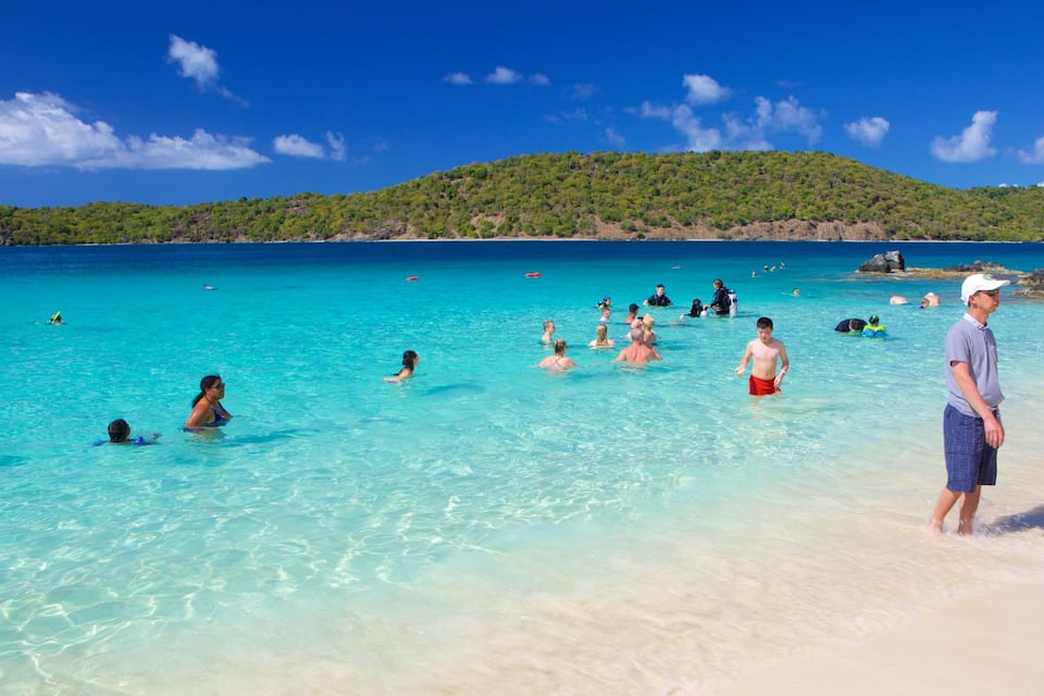 Een strand met helder turquoise water, mensen die zwemmen en zonnebaden, en een heuvel met begroeiing op de achtergrond.