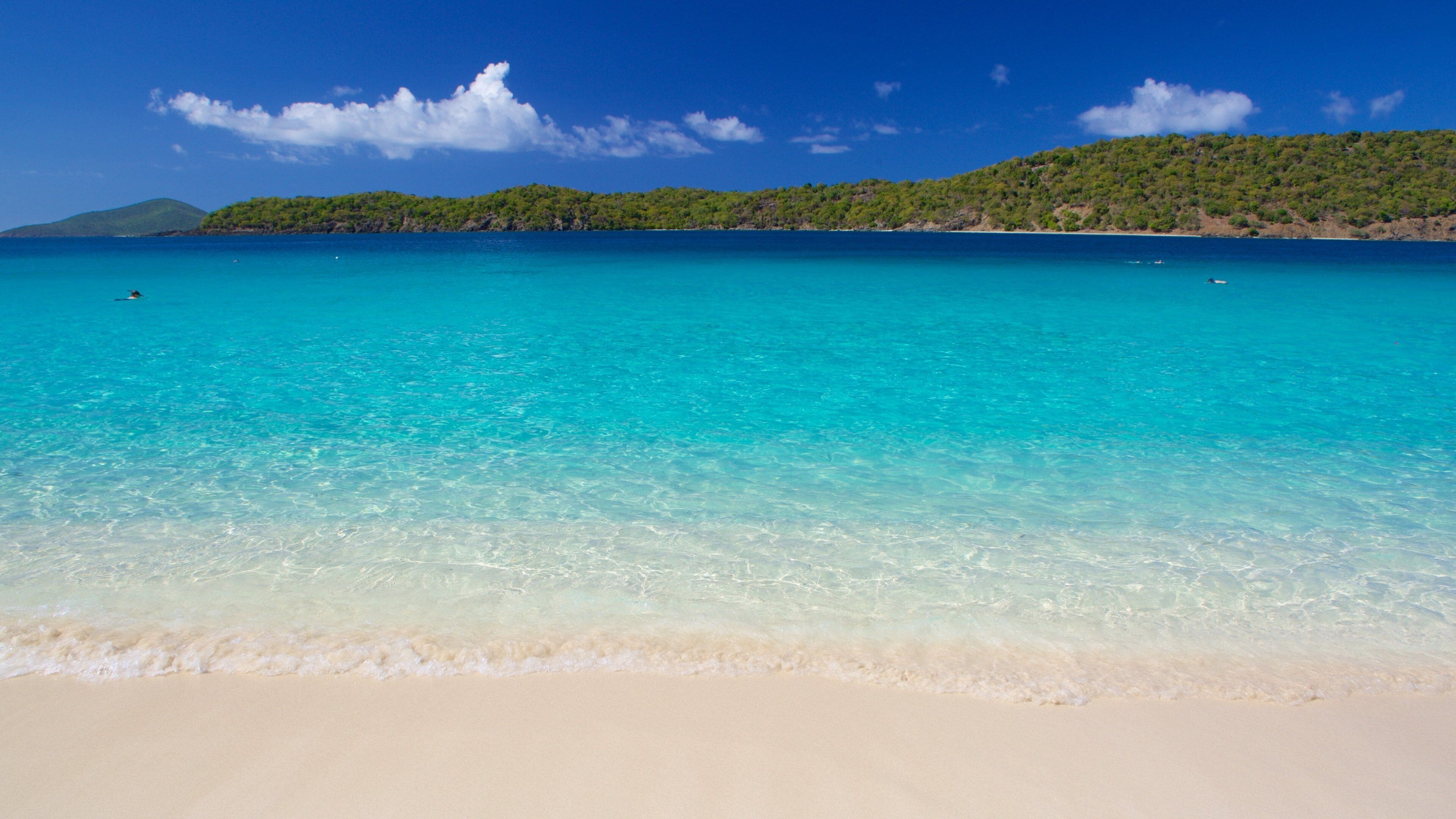 Ein Strand mit klarem türkisblauem Wasser, sandigem Ufer und einem grünen Hügel im Hintergrund.