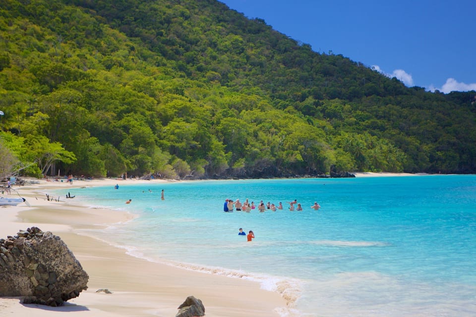 A beach with clear turquoise water, people swimming, and a forested hill in the background.