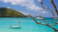 Une mer turquoise où l’on aperçoit une personne pratiquant le paddle, un voilier et un petit bateau à moteur. Au loin, on distingue une colline avec des bâtiments et un ciel dégagé.
