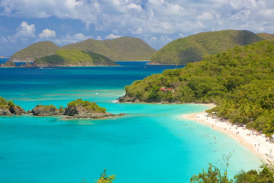 Une plage tropicale aux eaux turquoise et limpides, bordée de collines verdoyantes et surmontée d’un petit bâtiment rouge.