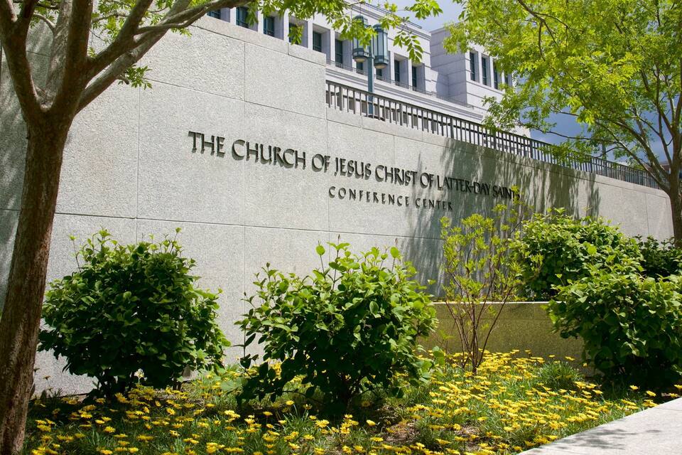 A building with a sign reading "The Church of Jesus Christ of Latter-day Saints Conference Center" surrounded by greenery and yellow flowers.