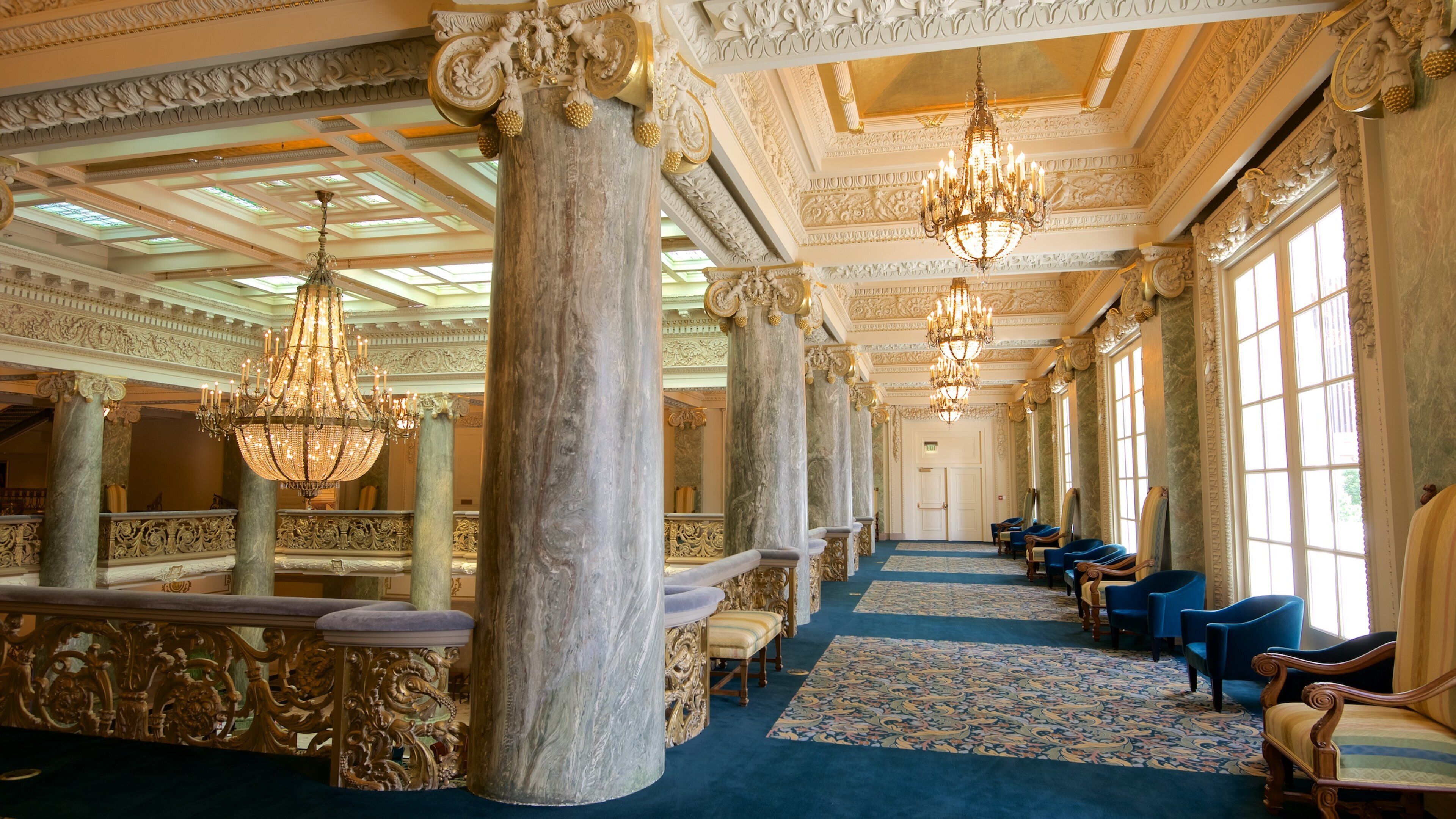 An opulent hall with ornate columns, chandeliers, and blue carpeting.