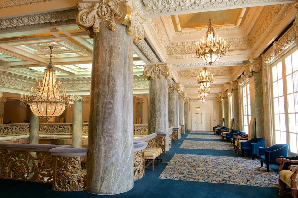 An opulent hall with ornate columns, chandeliers, and blue carpeting.