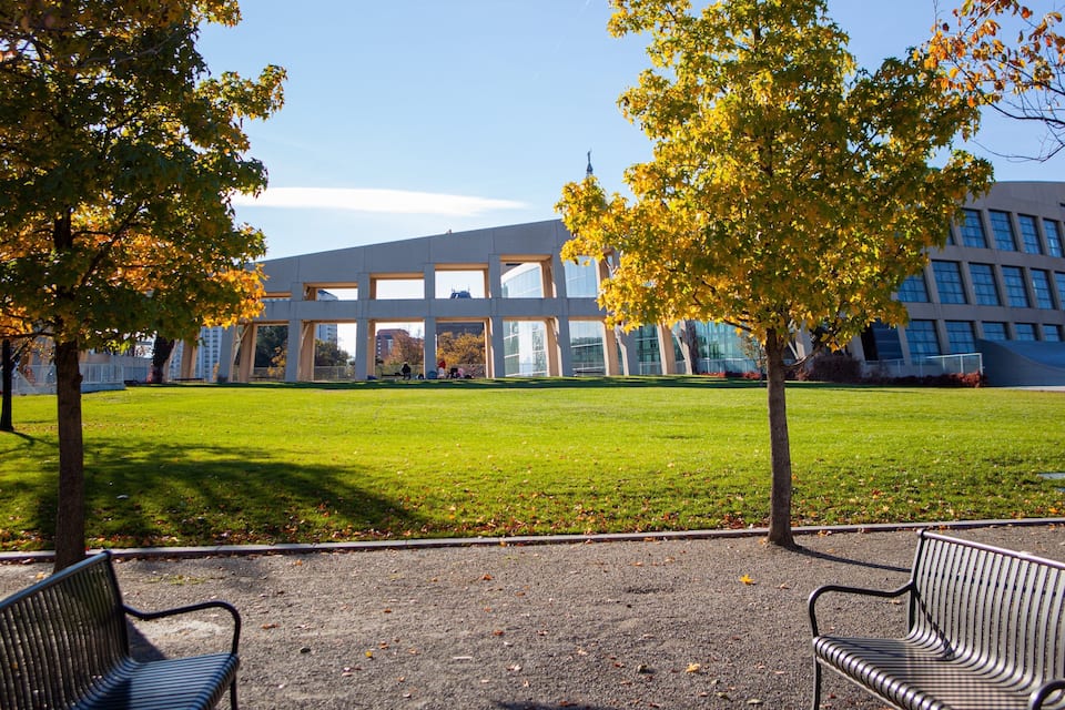 A modern building with a glass facade, surrounded by a grassy area with benches and trees with autumn leaves.