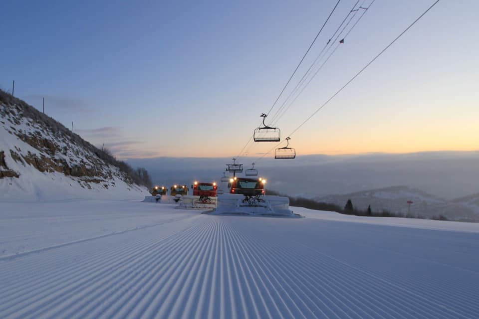 Pista de esquí nevada con un telesilla y vehículos a lo lejos.